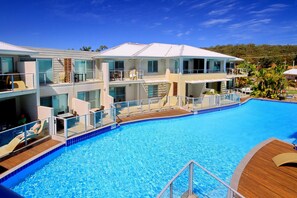A modern hotel with a large swimming pool, multiple balconies, and a view of the surrounding landscape.