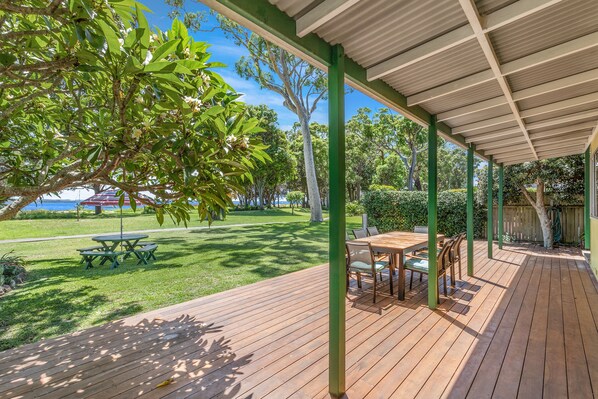 A covered outdoor deck with a table and chairs, surrounded by greenery and trees.