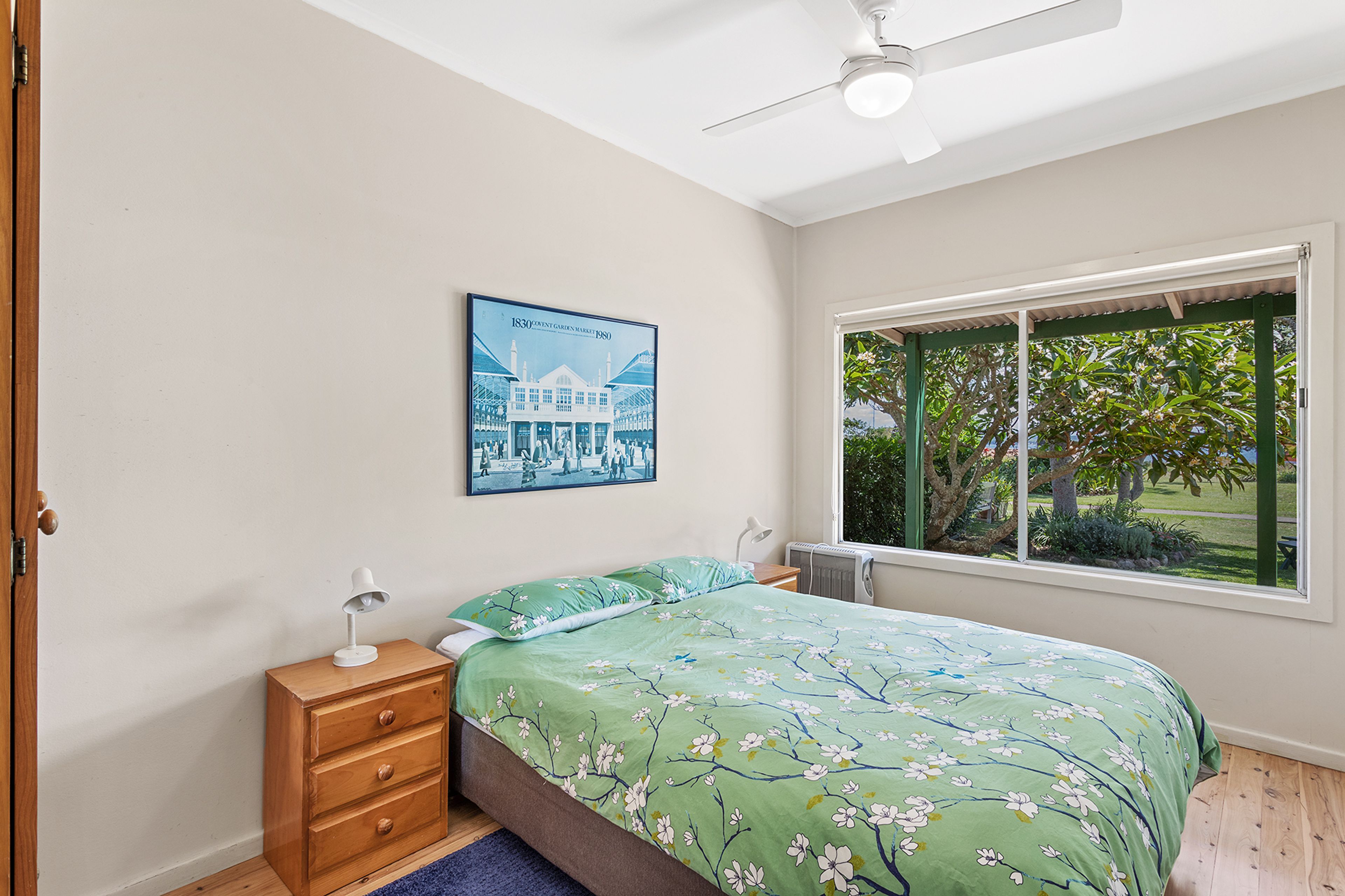 Une chambre à coucher comprenant un lit, une table de chevet, un bureau, une chaise, une fenêtre donnant sur un paysage verdoyant et un ventilateur de plafond.
