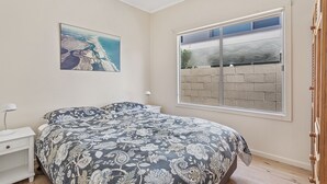 Quarto com cama, mesa de cabeceira, luminária, janela com vista para um muro de tijolos e um quadro na parede.