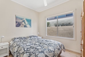 Een slaapkamer met een bed, nachtkastje, lamp, raam met uitzicht op een bakstenen muur, en een schilderij aan de muur.