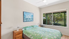 A bedroom with a bed, a nightstand, a desk lamp, a radiator, a window with a view of trees and a building, and a framed picture on the wall.