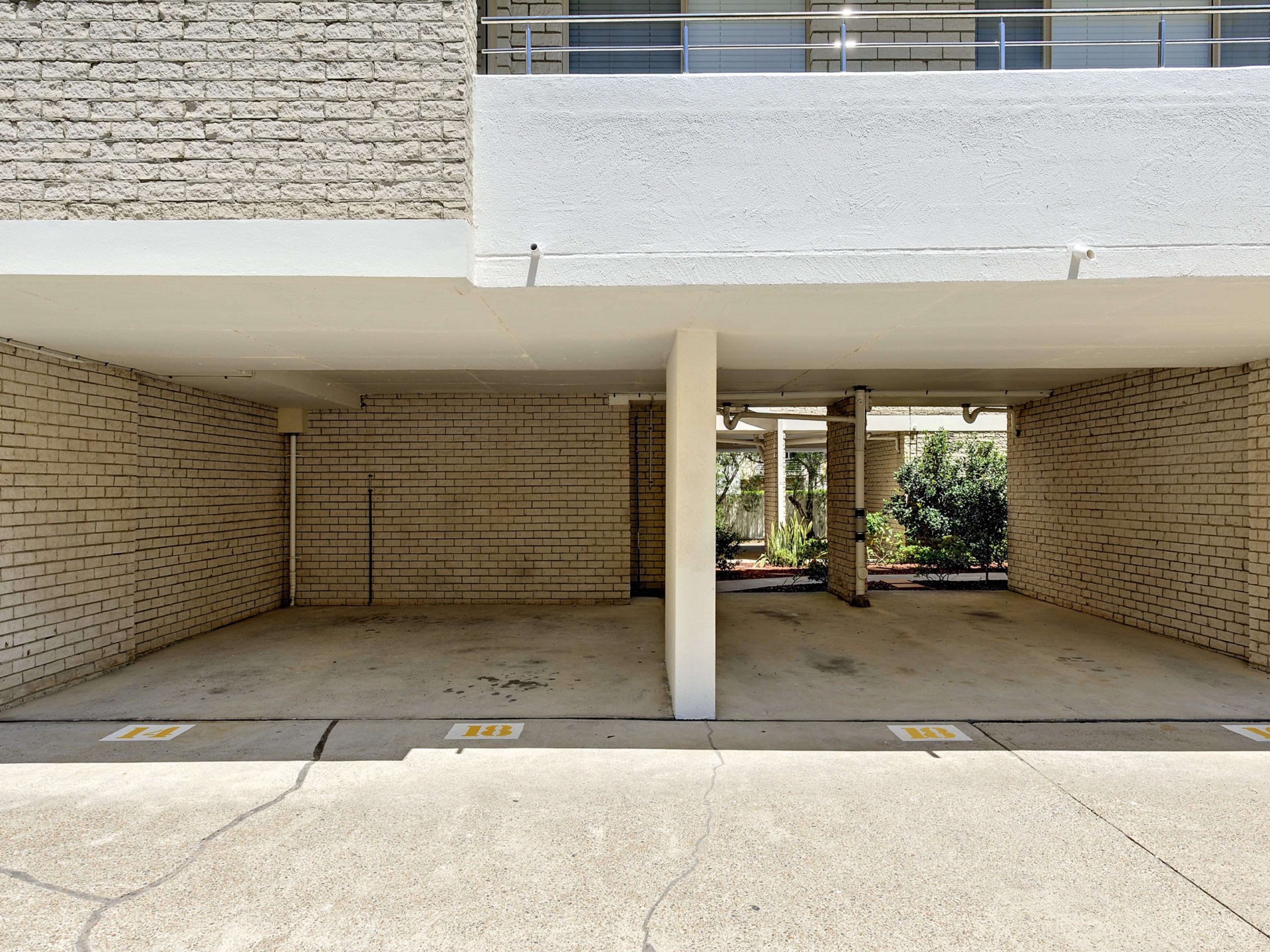 An empty parking area with brick walls and concrete flooring, featuring a white column structure and a visible outdoor area with greenery.