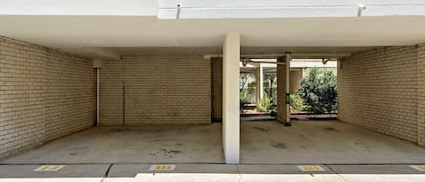 En tom parkeringsplats med tegelväggar och betonggolv, med en vit kolonnstruktur och ett synligt utomhusområde med grönska.