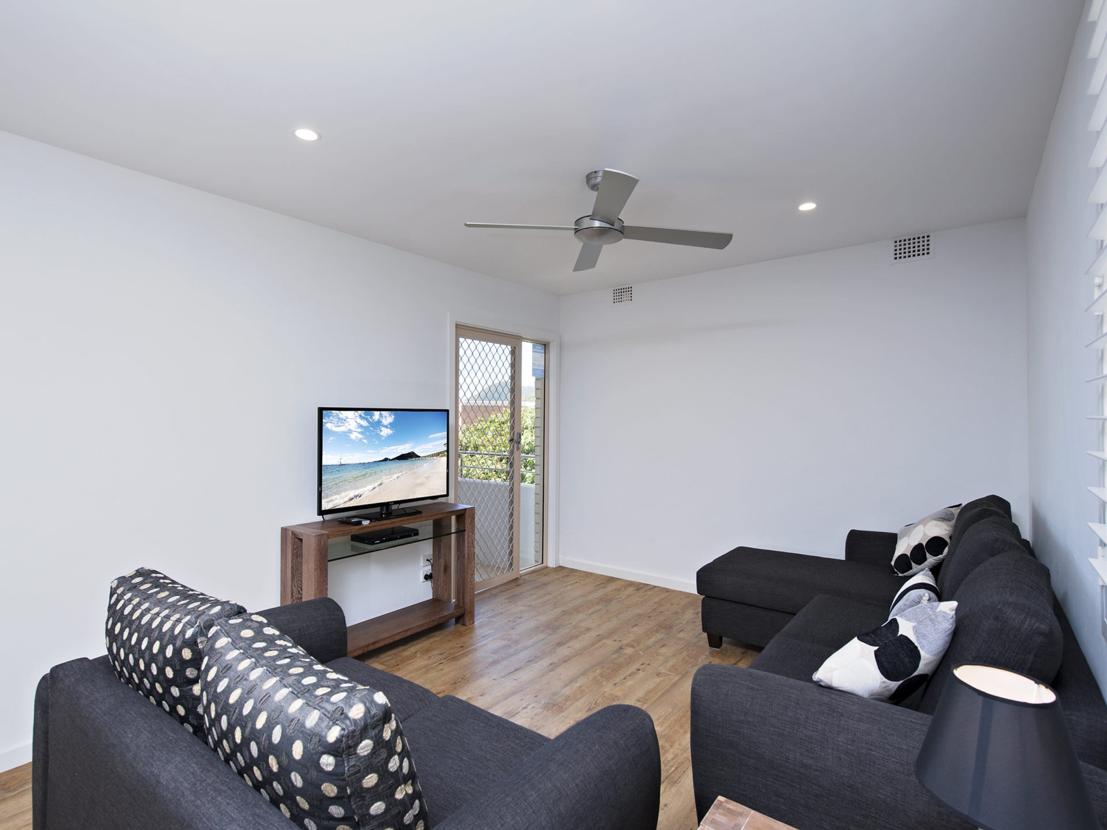 A modern living room with a flat-screen TV on a wooden stand, a ceiling fan, and a large window with a view of the outdoors.