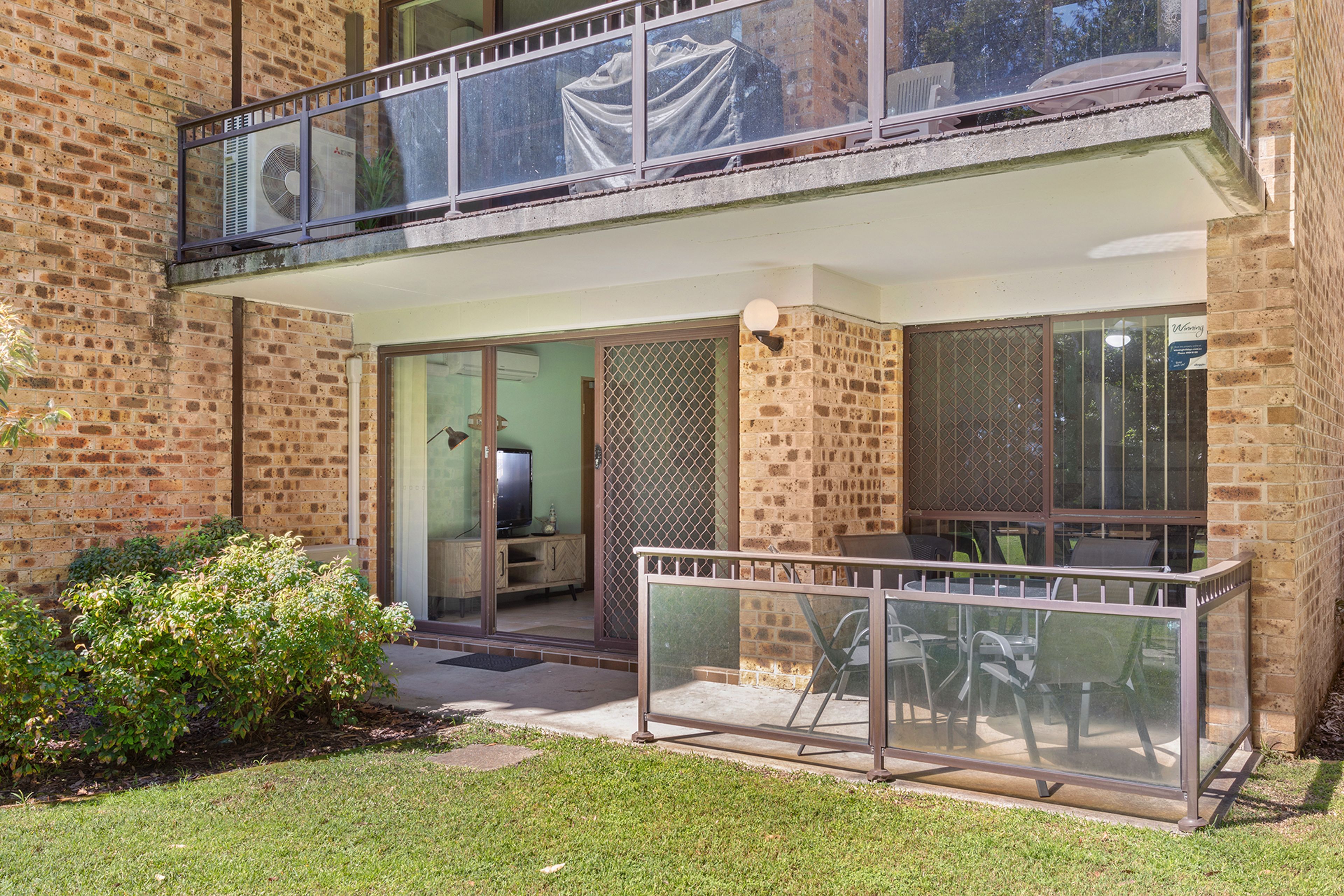 A two-story building with a balcony, a sliding glass door leading to a room with a television, and a small patio area with outdoor furniture.
