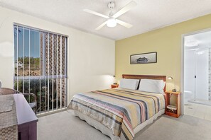 Quarto com cama, mesa de cabeceira, relógio na parede, ventilador de teto, janela com vista e porta.