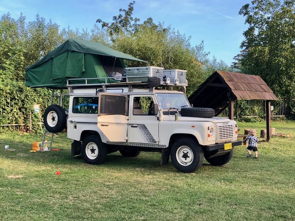 Un SUV blanc, équipé d’une tente de toit et d’un pneu de secours fixé à l’arrière, est garé sur de l’herbe, avec une structure en bois en arrière-plan.
