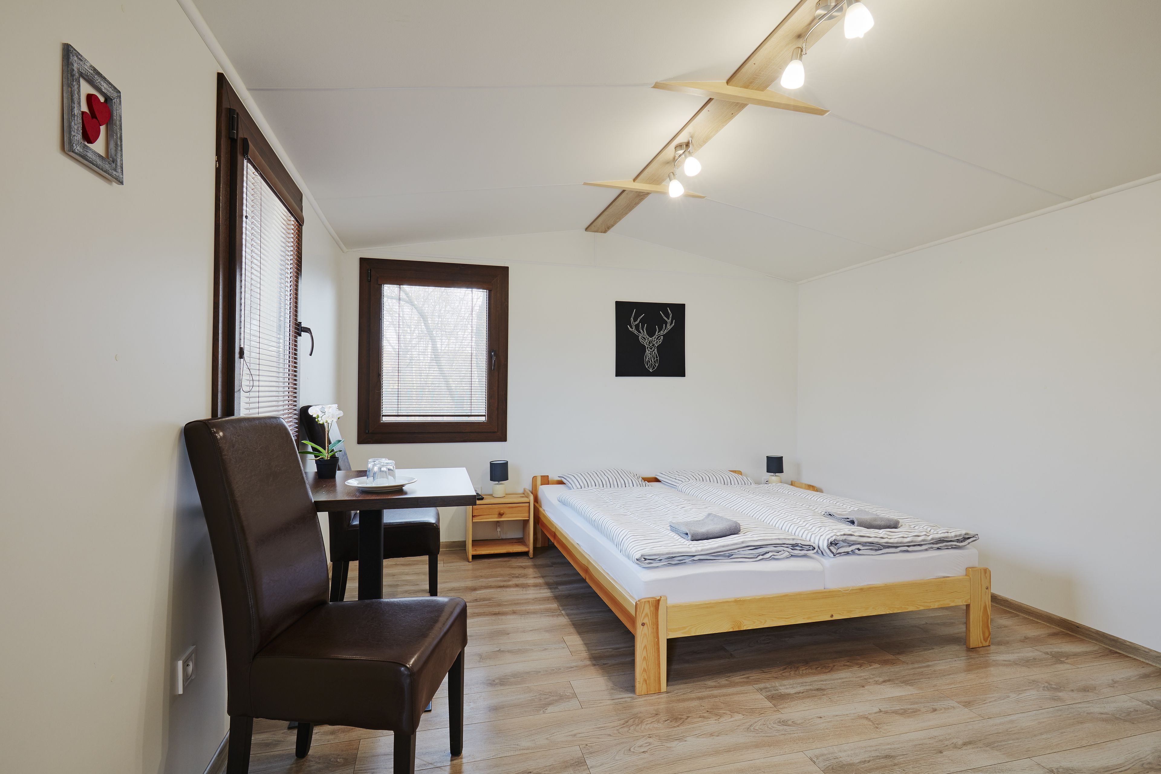 Een slaapkamer met een houten bed, een bureau met stoel, een klein tafeltje en een raam met luxaflex.