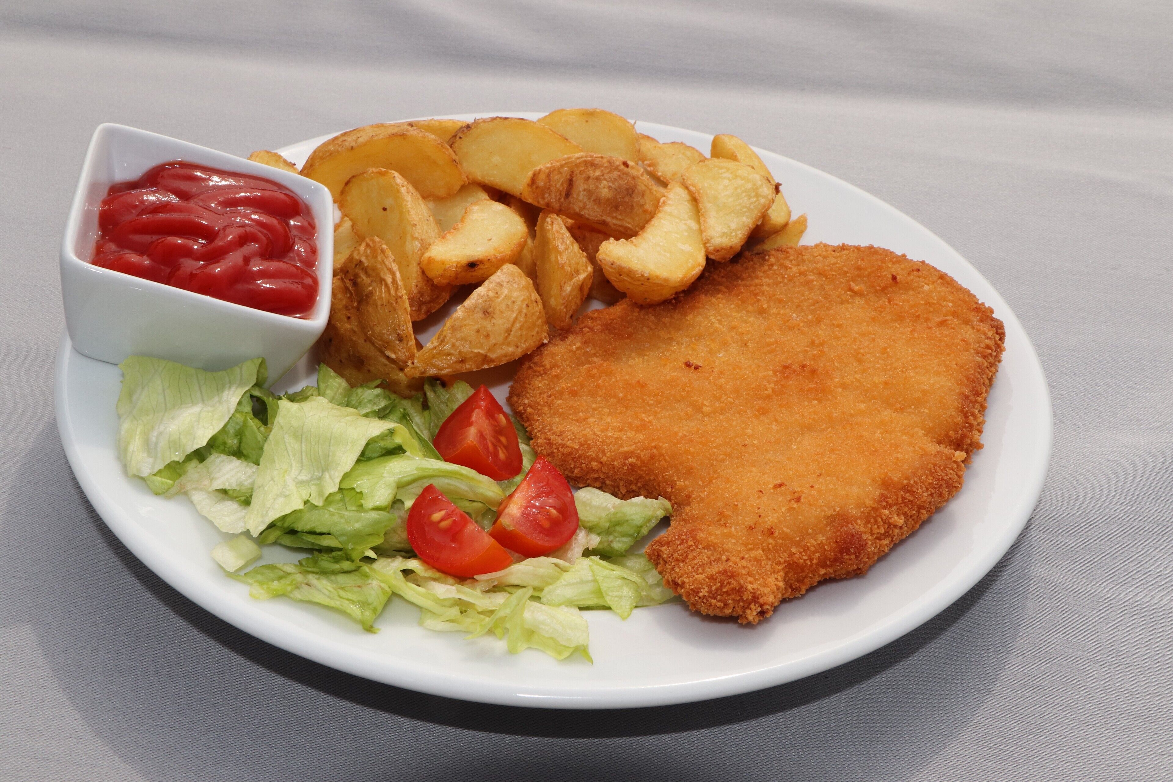 Une assiette contenant une escalope panée, des quartiers de pommes de terre, de la laitue et un petit bol de ketchup.