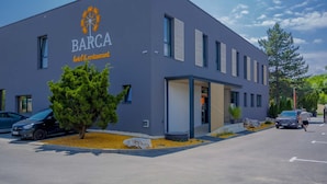 A modern building with a sign reading "BARCA hotel & restaurant" and a logo above it. There are cars parked in front and a tree to the left. - Hotel & Restaurant Resort Barca  - Campsite (Kosice)