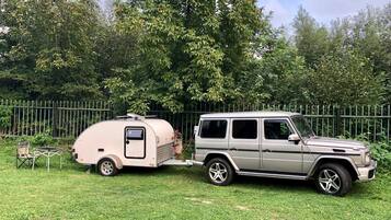 Un SUV color beige remolca un pequeño remolque con un panel solar en la parte superior, estacionado en un área cubierta de césped con una cerca metálica y árboles al fondo.