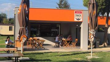Un espace extérieur aménagé avec des tables et des chaises, un panneau indiquant un bar à collations et des parasols.