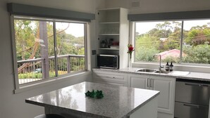 Una cocina moderna con una isla central, armarios blancos, electrodomésticos de acero inoxidable y grandes ventanales que ofrecen vistas a zonas verdes y un parque infantil.