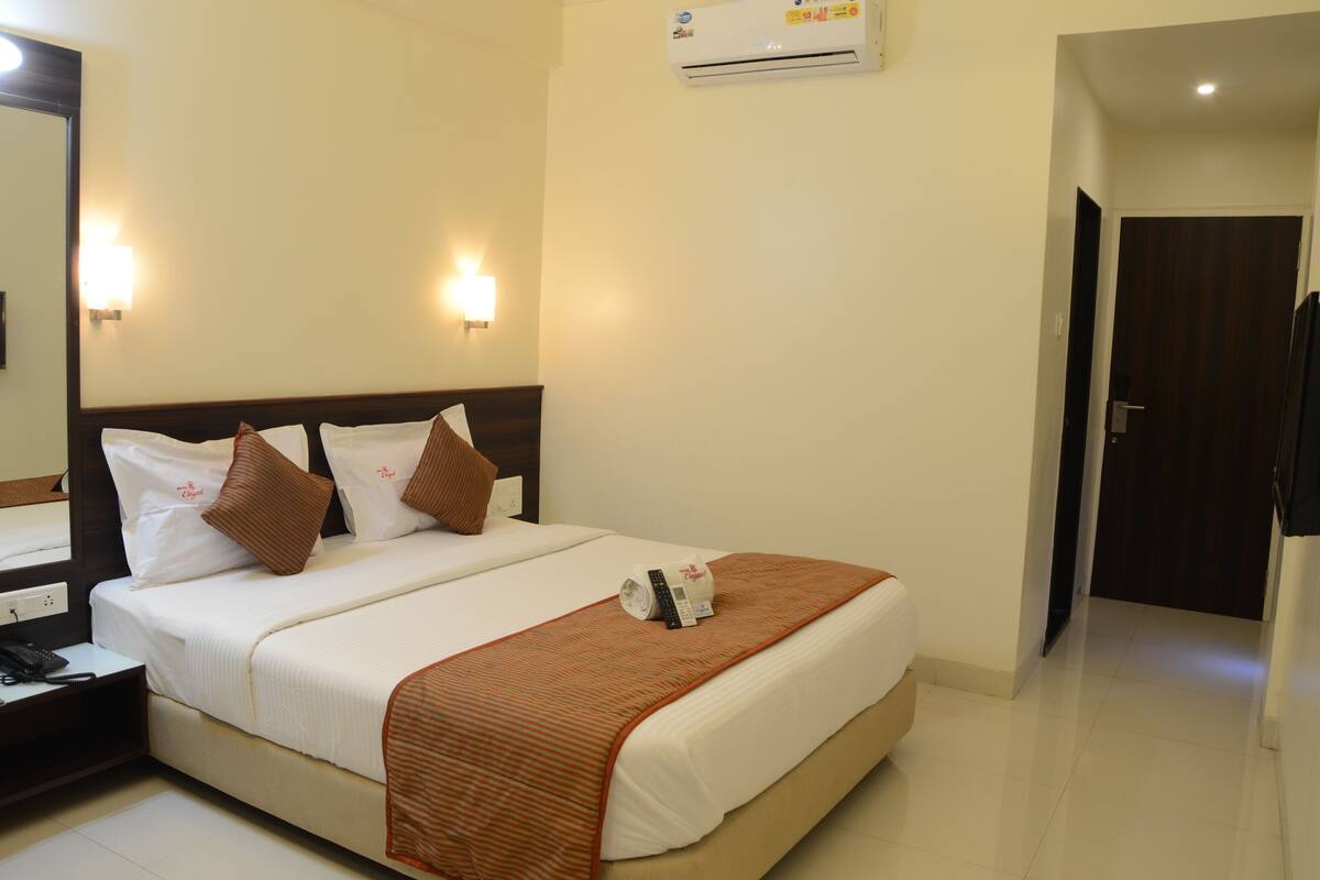 A hotel room with a bed, bedside table, lamp, and an air conditioning unit.