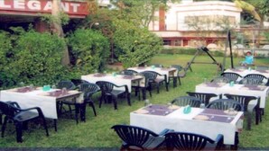 Un espace extérieur aménagé pour les repas, avec des tables et des chaises, une balançoire et un bâtiment dont on aperçoit une enseigne à l’arrière-plan.