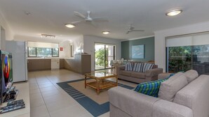 Ein modernes Wohnzimmer mit einer Couch, einem Couchtisch und einem Fernseher. Es gibt eine Küchenzeile mit Kühlschrank, Spüle und einem Fenster. Der Raum verfügt über Deckenventilatoren und Einbaustrahler.