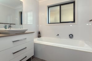 A modern bathroom with a freestanding bathtub, a large mirror, and a window above the tub. - Beach House Family Friendly in Fingal Bay (Fingal Bay)