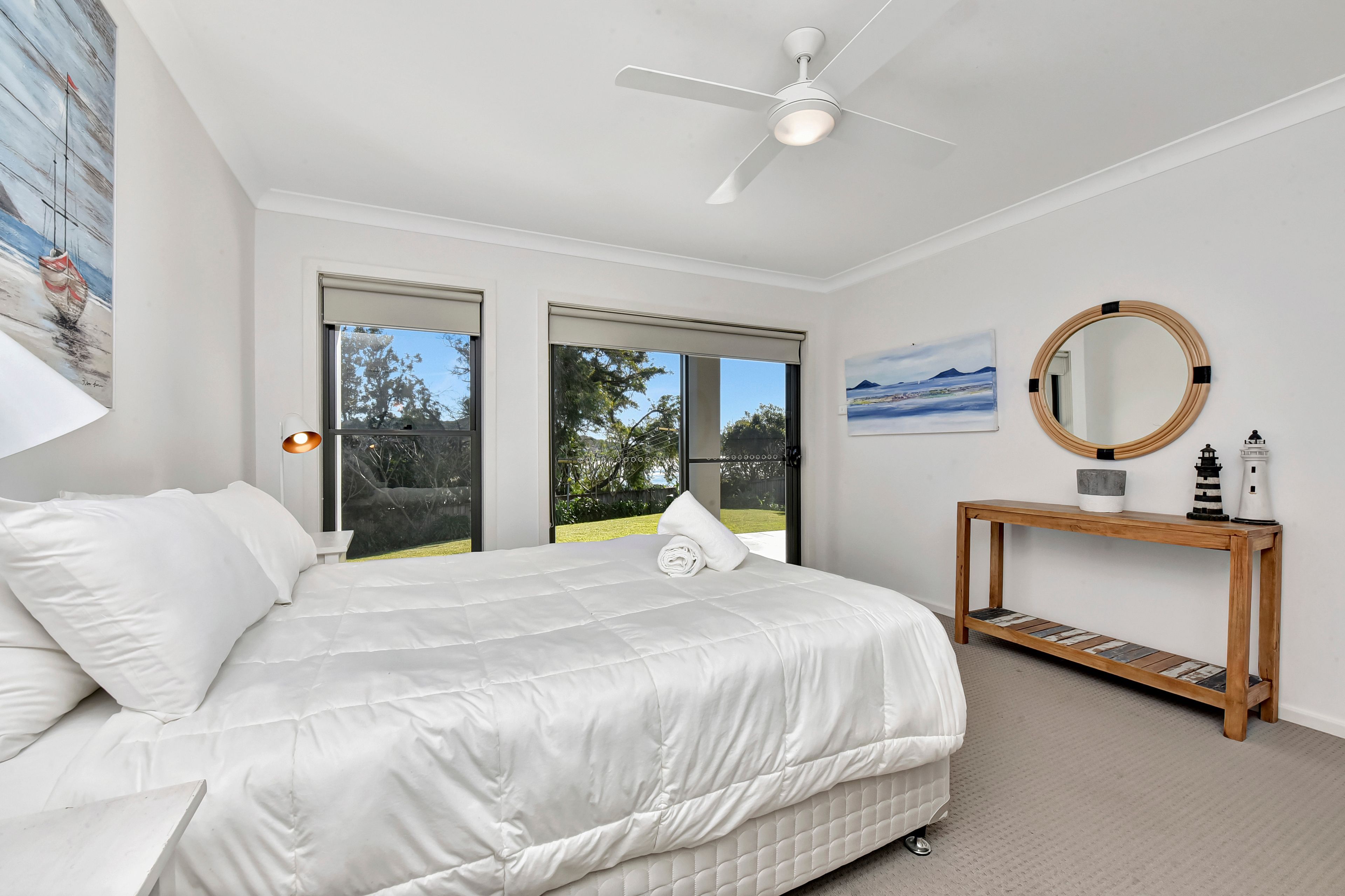 A bedroom with a bed, a ceiling fan, a large window, and a bedside table with a mirror.