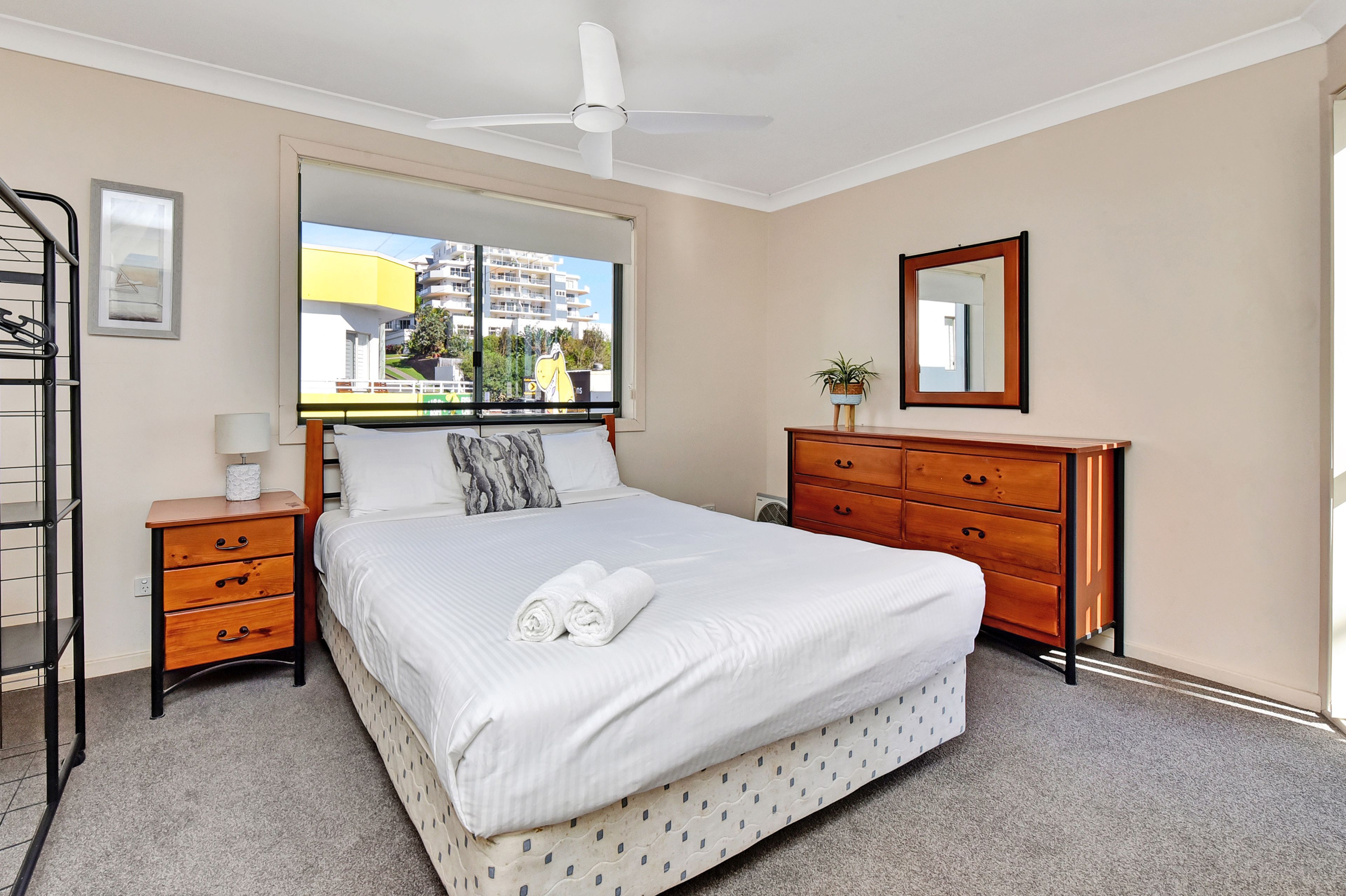 A hotel room with a bed, a dresser, a mirror, a window with a view of buildings, and a ceiling fan.