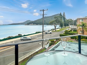 A balcony with a clear view of a coastal road, a parked car, and a table with two empty wine glasses. - Bella Vista 3 Shoal Bay Apartment with Water Views (Shoal Bay)