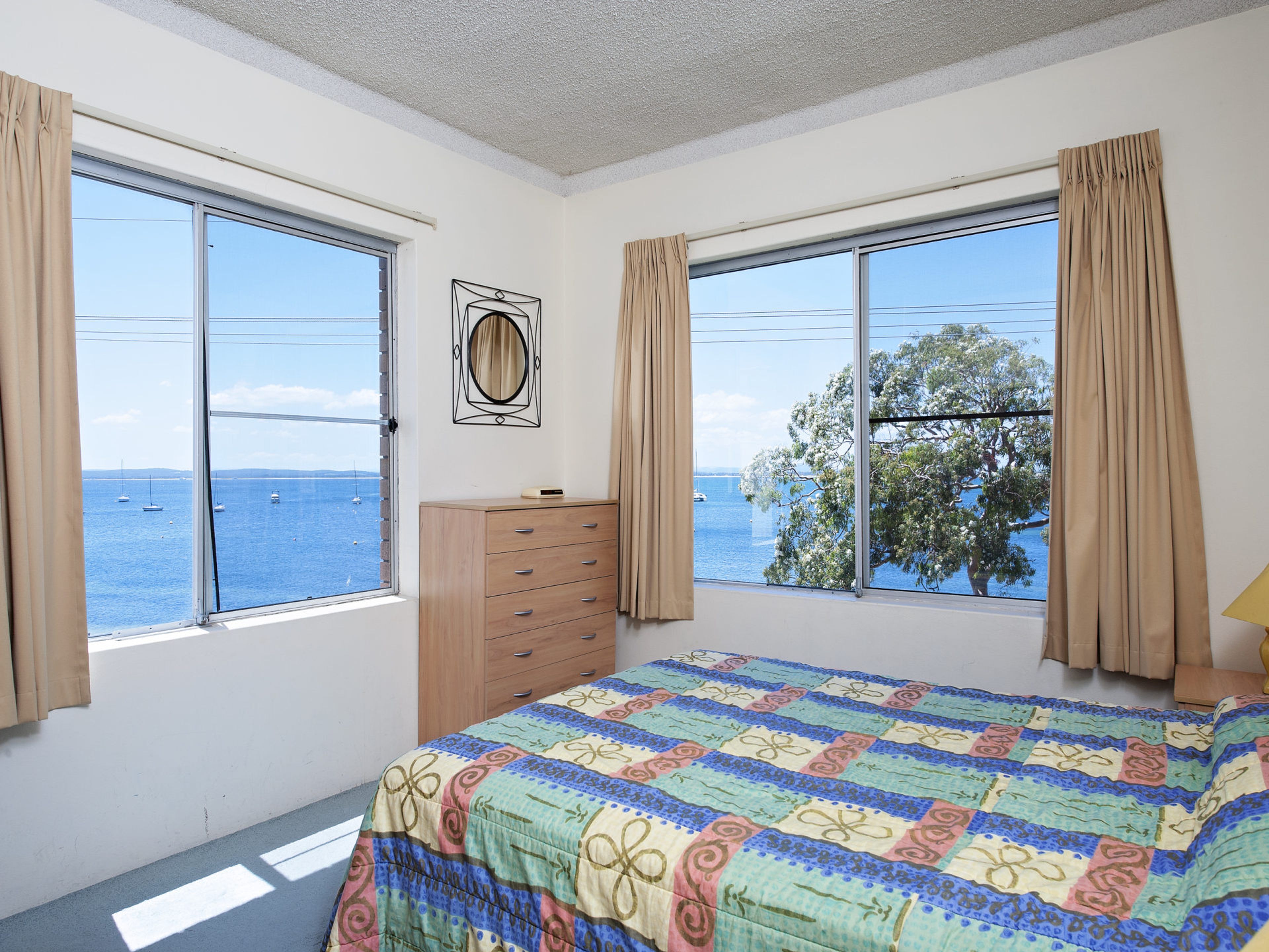 A bedroom with a large window, a bed with a colorful quilt, a wooden dresser, and a wall-mounted mirror.