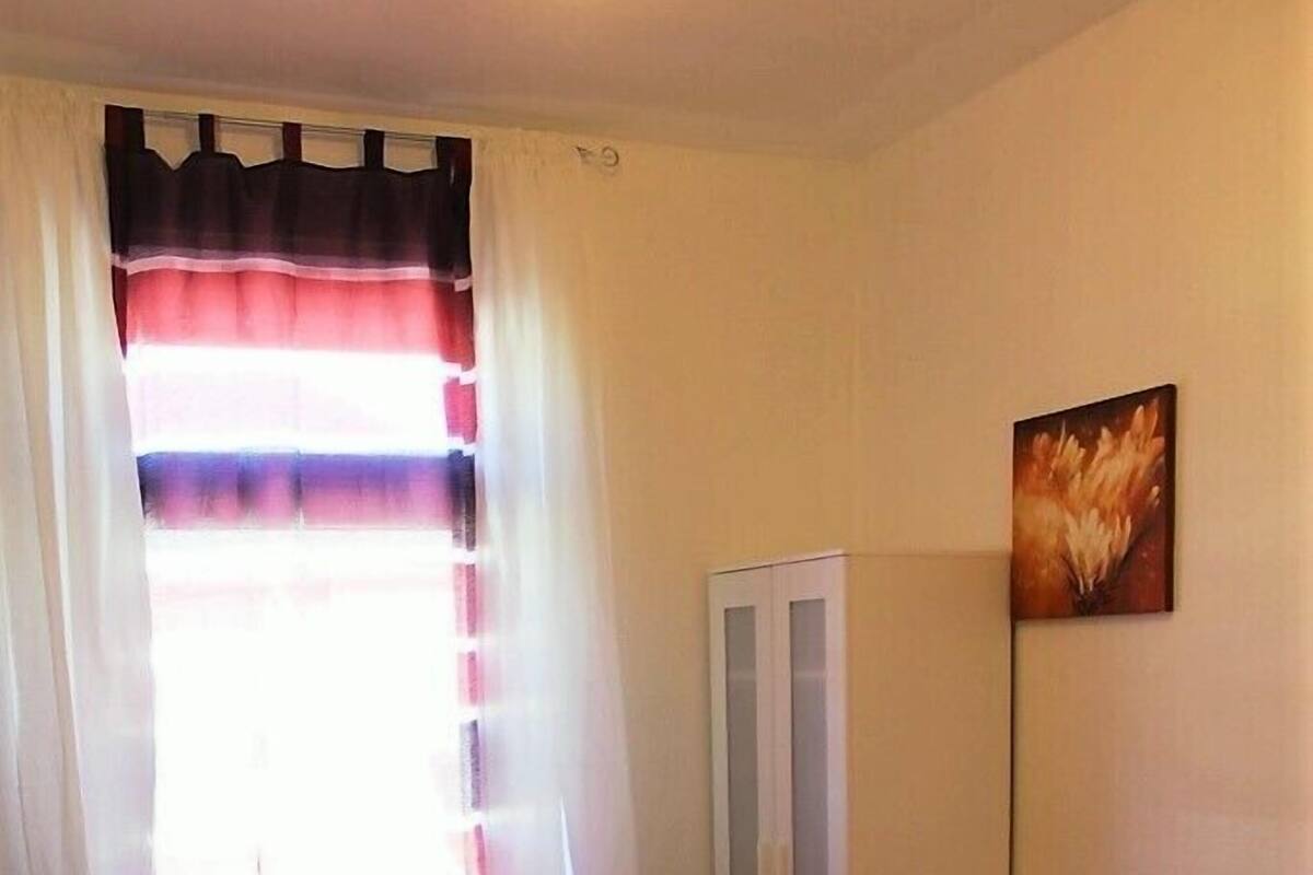 Una habitación pequeña con cama, escritorio, silla, armario y ventana con cortinas.