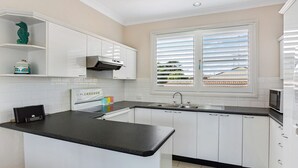 A modern kitchen with white cabinets, a black countertop, and a window with blinds. - Sandy Shores at Shoal Bay (Shoal Bay)