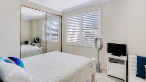 A bedroom with a bed, a TV, a mirror, and a window with blinds. - Sandy Shores 1 Pet Friendly Shoal Bay Home (Shoal Bay)