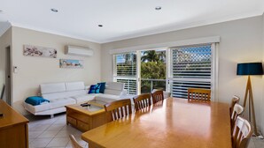 A modern living room with a white sofa, wooden dining table, and chairs. There is a large window with blinds, a ceiling fan, and artwork on the walls. - Sandy Shores at Shoal Bay (Shoal Bay)