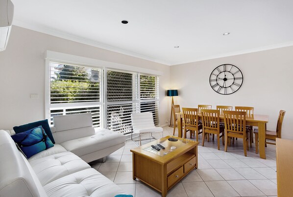A living room with a white sofa, a coffee table, a dining table with chairs, and a wall clock.