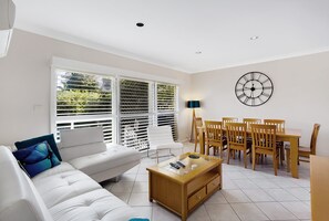 A living room with a white sofa, a coffee table, a dining table with chairs, and a wall clock.
