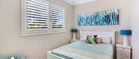 Ein Schlafzimmer mit einem Bett, zwei Nachttischen, einem Fenster mit Jalousien und einem an der Wand hängenden abstrakten Gemälde.