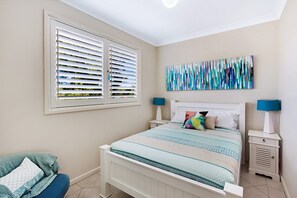 A bedroom with a bed, two bedside tables, a window with blinds, and a wall-mounted abstract painting.
