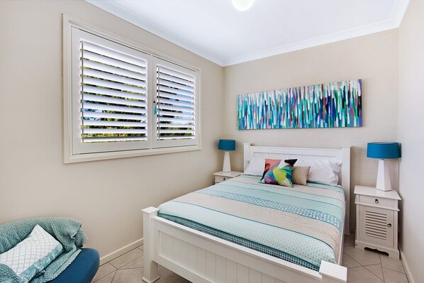 A bedroom with a bed, two bedside tables, a window with blinds, and a wall-mounted abstract painting.