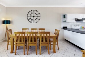 A dining area with a wooden table and chairs, a wall-mounted clock, and a kitchen with white cabinets and a black countertop.