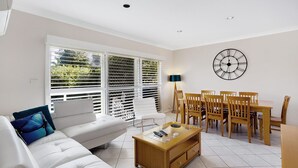 A modern living room with a white sofa, a wooden coffee table, and a dining area with wooden chairs and a table. - Sandy Shores at Shoal Bay (Shoal Bay)