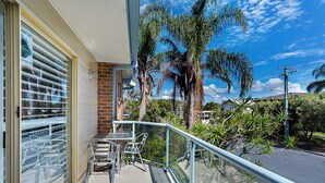 A balcony with a table and chairs, overlooking a street with palm trees and a clear sky. - Sandy Shores 1 Pet Friendly Shoal Bay Home (Shoal Bay)