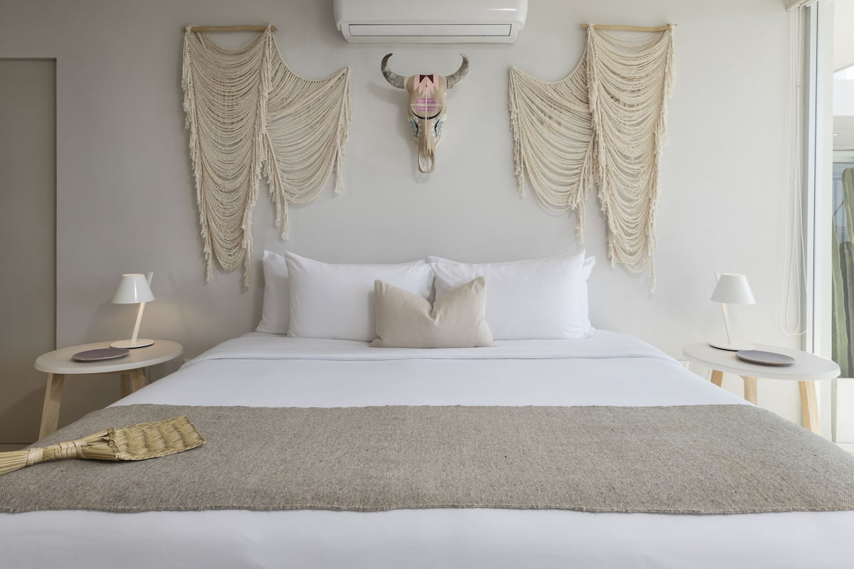 A neatly made bed with white linens, a decorative skull wall hanging, and two bedside tables with lamps.
