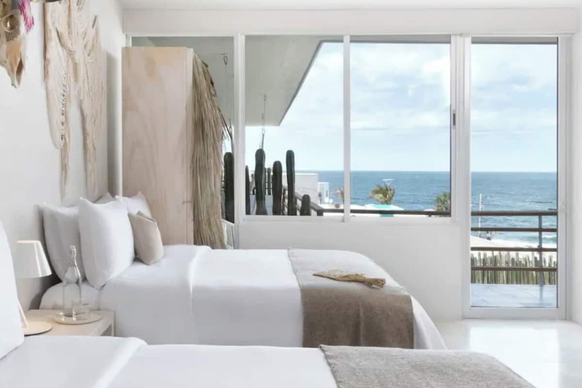 A modern hotel room with two beds, a large window with a view of the sea, and a balcony with a railing.