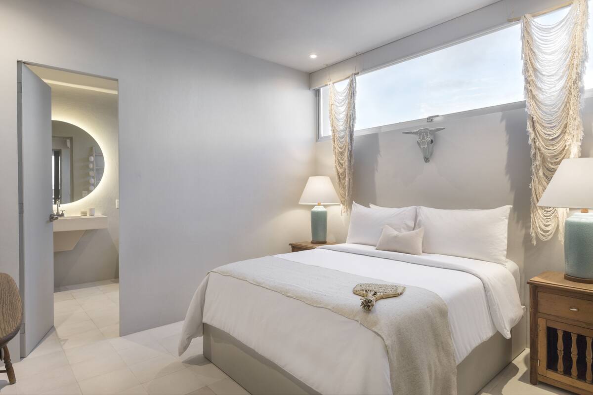 A modern bedroom with a large bed, bedside lamps, a mirror, and a decorative wall piece.