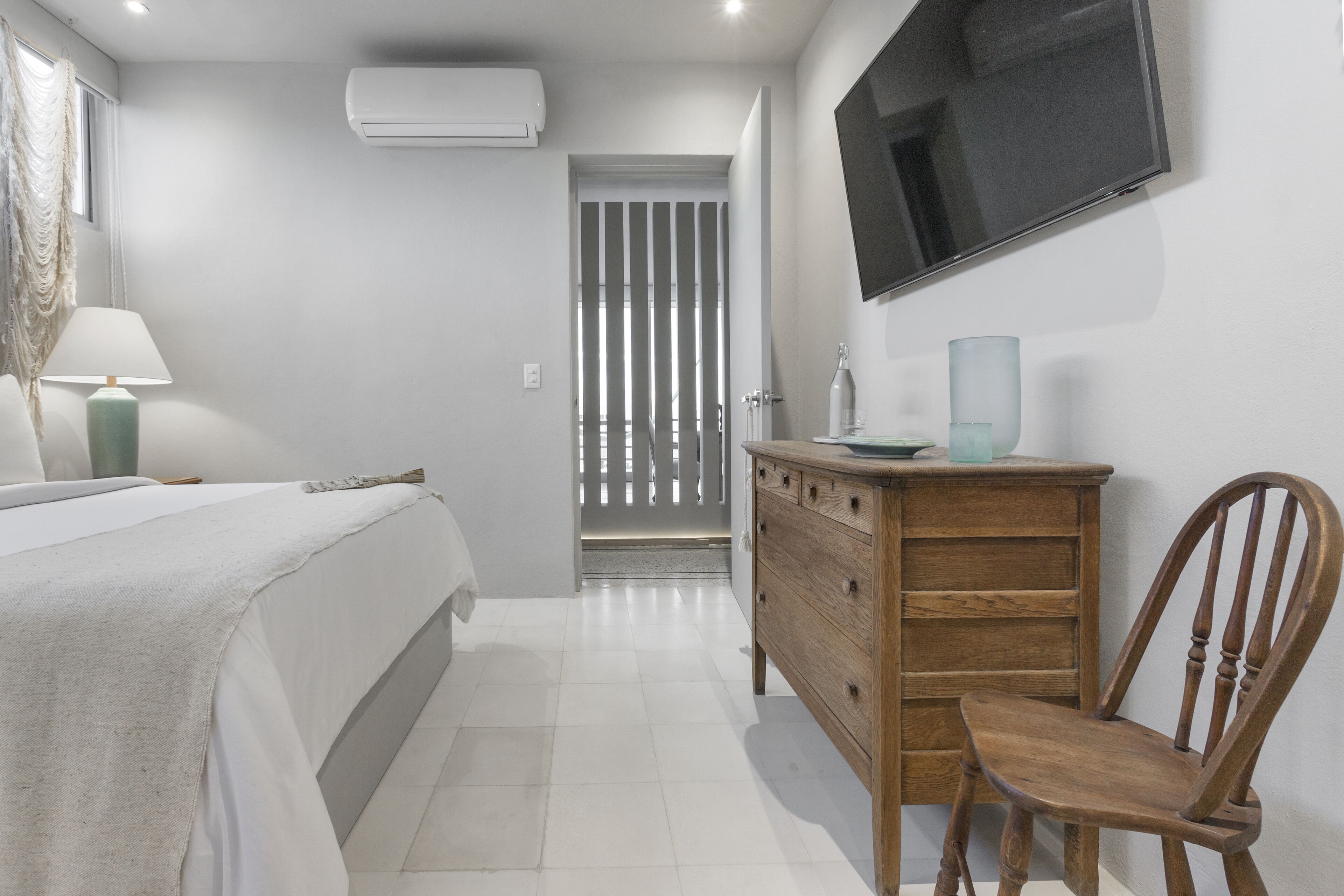 Un dormitorio con una cama, un tocador de madera, una silla, un televisor montado en la pared y una puerta que da a otra habitación.