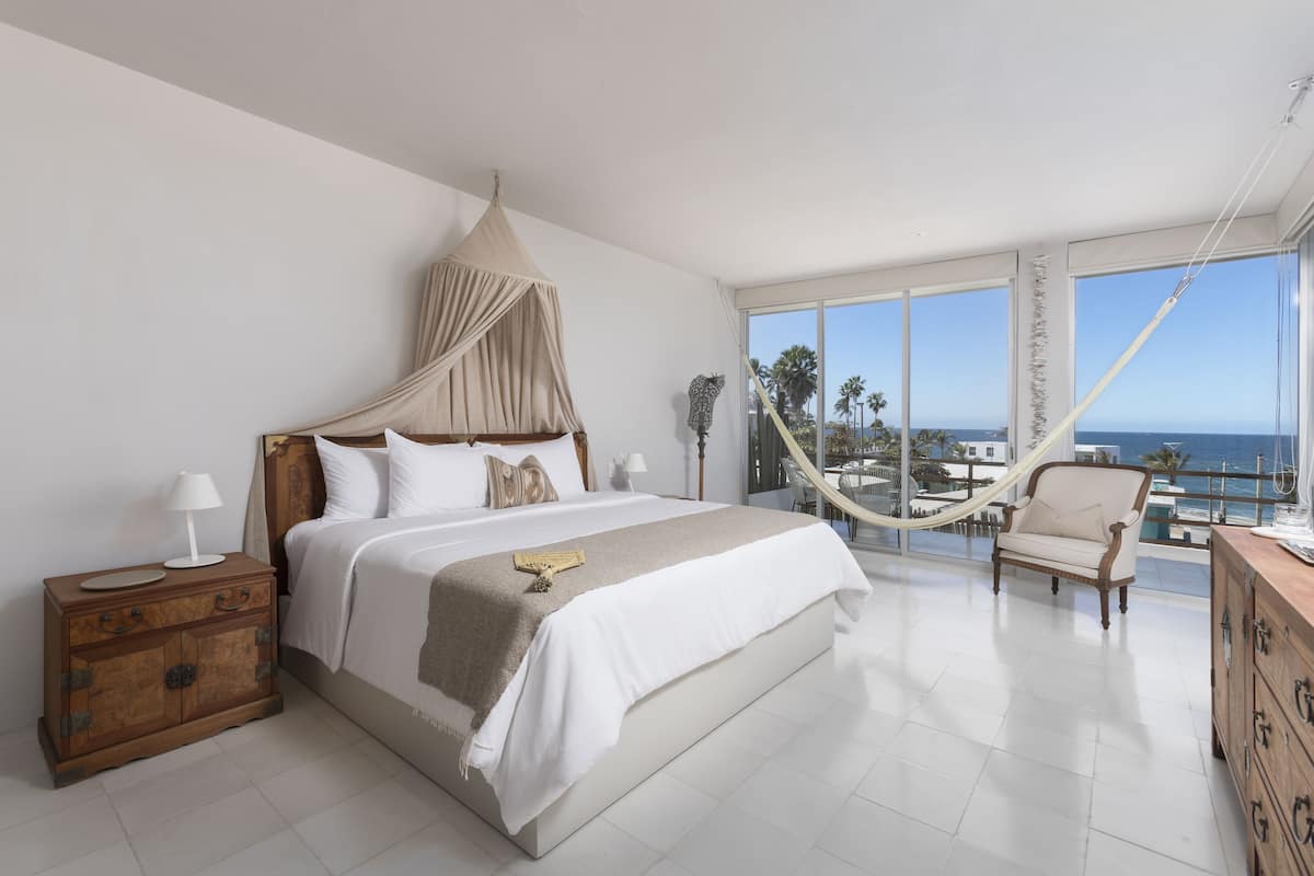 A spacious bedroom with a large bed, a wooden nightstand, a chair, and a view of the ocean through large windows.