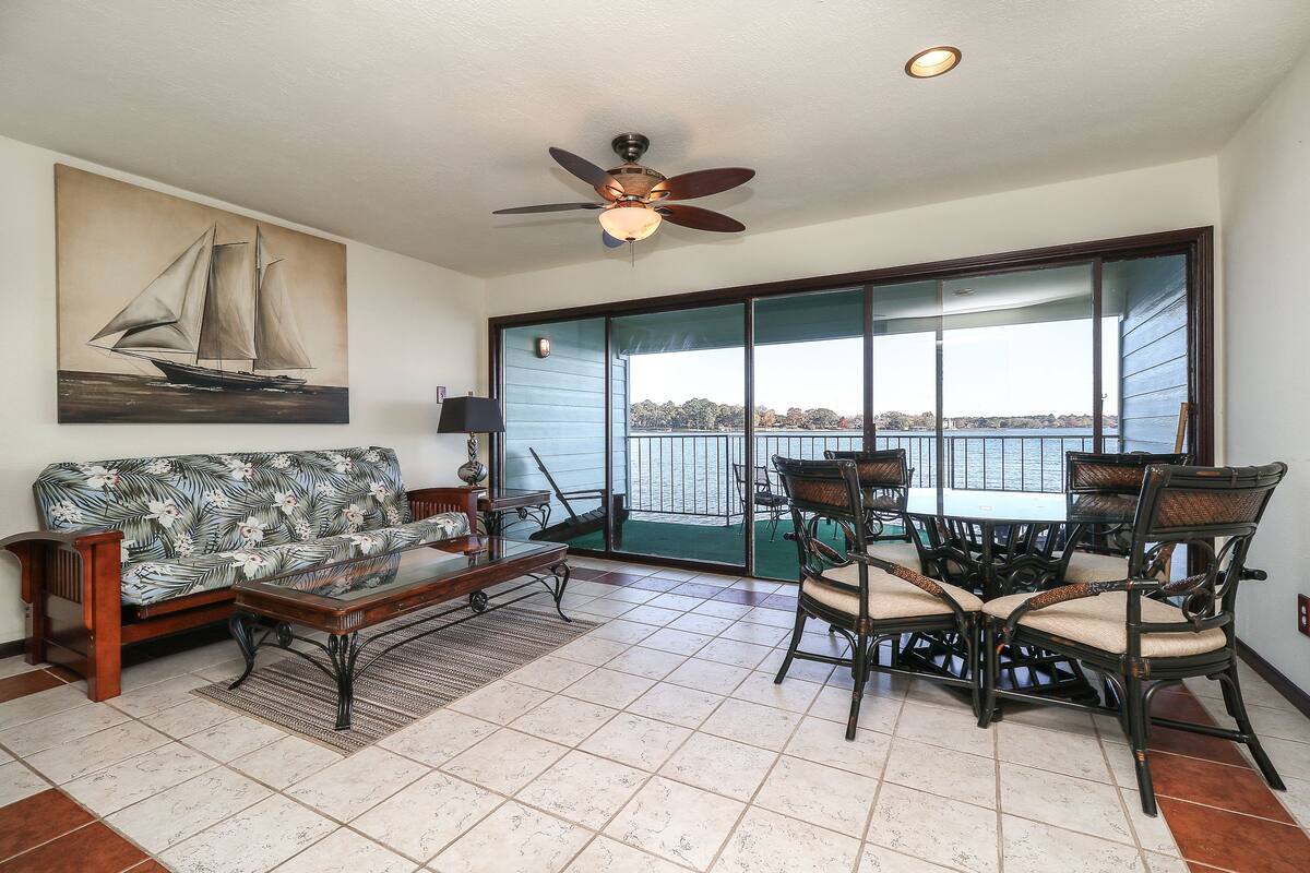 A living room with a sofa, coffee table, and dining set. There is a ceiling fan and a large window with a view of the water.