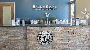 Ett välkomstområde med en vägg av sten, en logotyp och en skylt med texten ”Hand & Stone Massage and Facial Spa”.