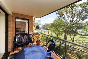 A balcony with a table and chairs, a potted plant, and a view of a tree-lined street.