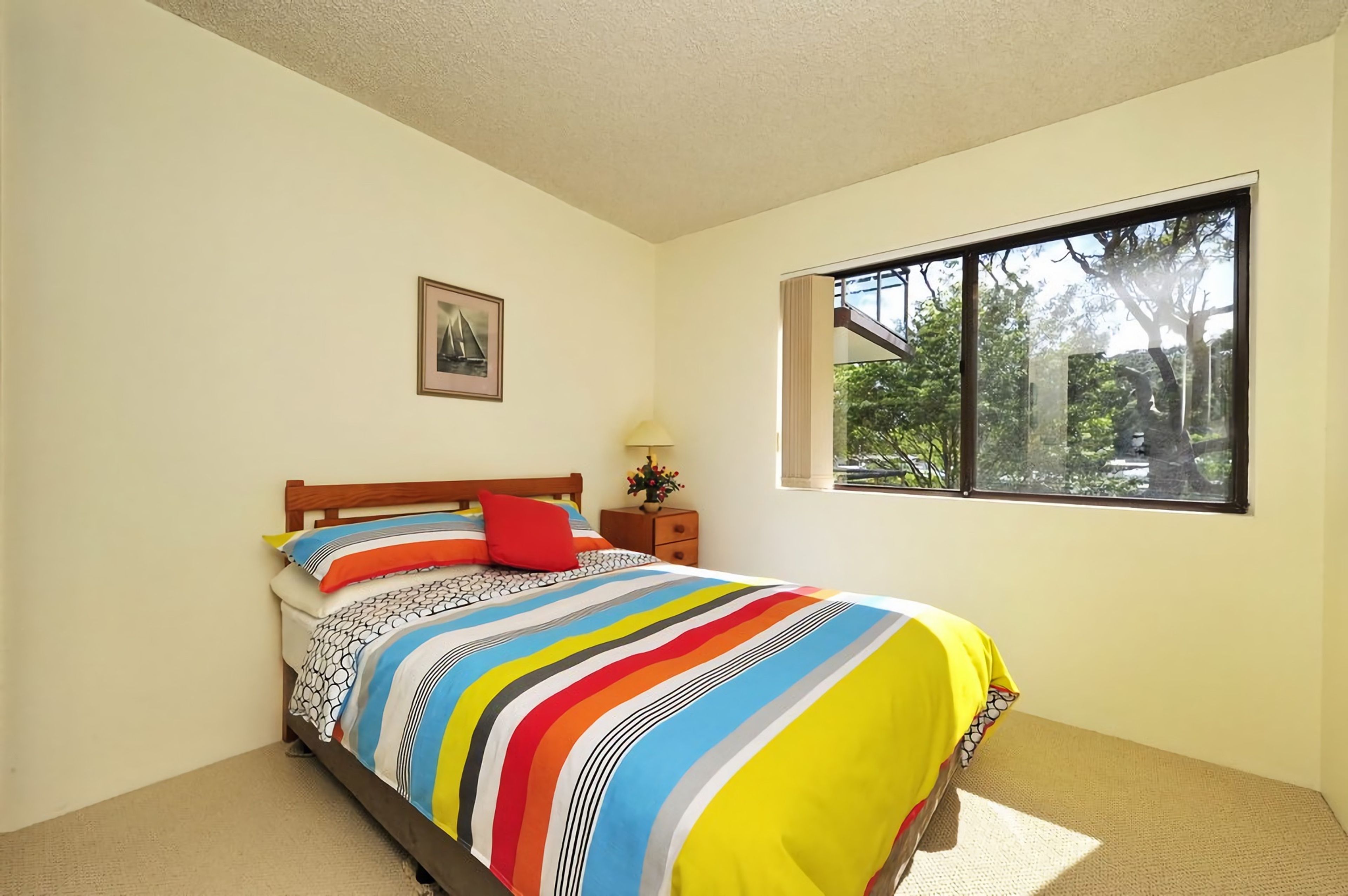 A bedroom with a bed, a nightstand, a lamp, a window, and a framed picture on the wall.