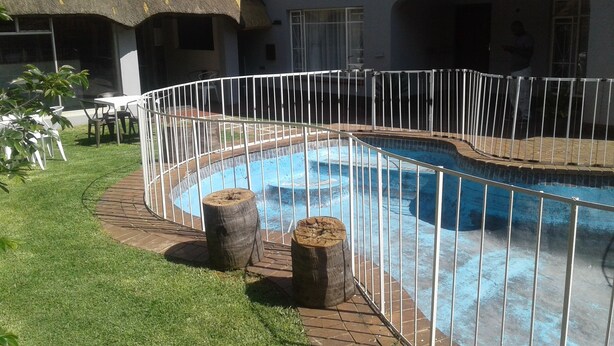 A swimming pool with a white metal fence, surrounded by a brick deck and grass. - Acacia Guesthouse (Kempton Park)