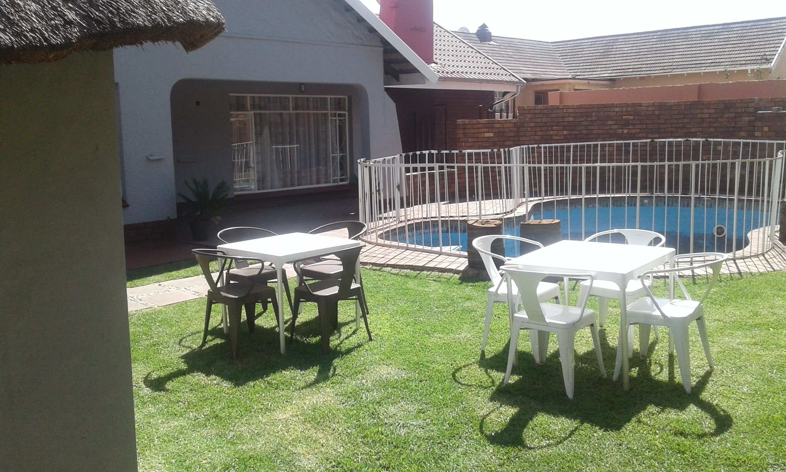 A backyard with a swimming pool, surrounded by a white metal fence, and a patio area with white tables and chairs.