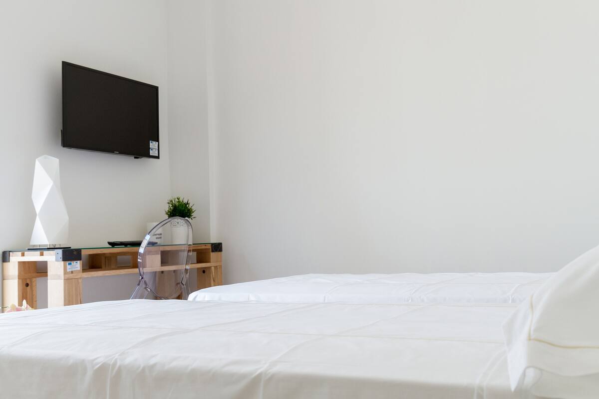A modern hotel room with a bed, a desk with a computer, a TV mounted on the wall, and a small plant on the desk.